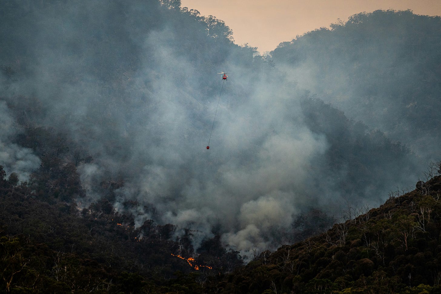 A hillside forest fire with a firefighting helicopter flying through the valley