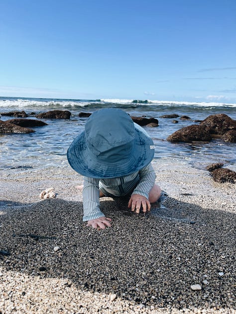 a toddler in a blue and white striped bathing suit and a blue hat plays in the sand on a beach.