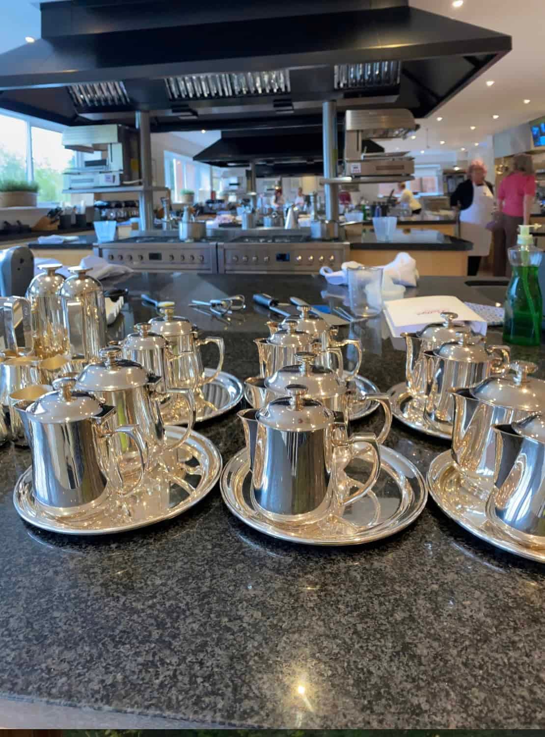 Bettys Cookery School kitchen, with rows of silver tea pots in the foreground. Collection of Silver Tea pots on a counter top.