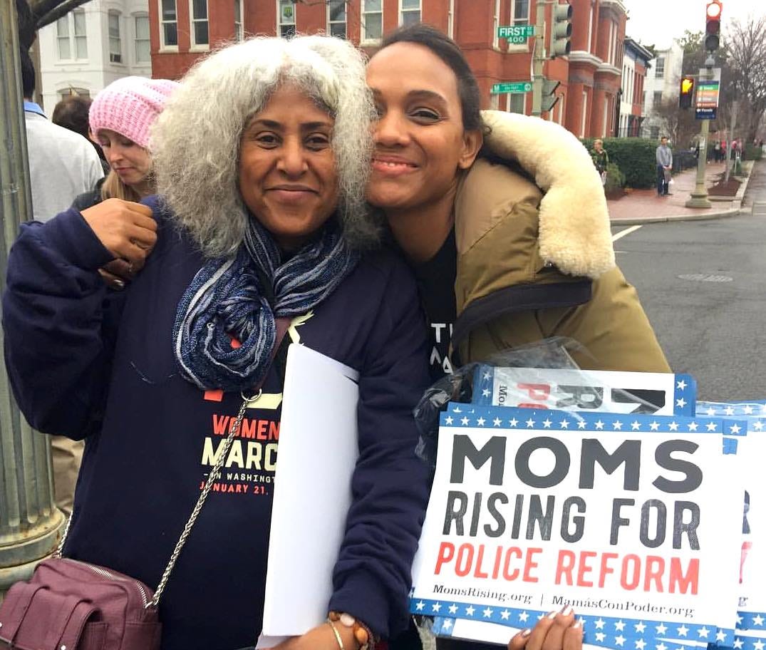 Makani standing with a young woman at a street protest, they hold signs that read "Moms Rising for Police Reform." They are dressed in winter coats and smiling.