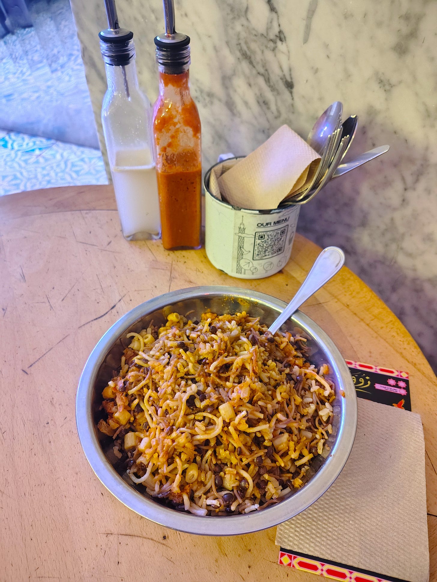 A metal bowl on a wooden round table filled with koshary and a spoon in it, with paper napkins on the side.