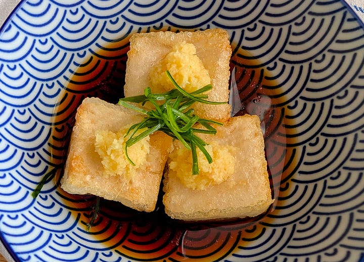 Agedashi Tofu in Light Soy-Based Sauce: Crispy, golden-brown cubes of agedashi tofu sit in a shallow pool of light, savory soy-based sauce. The dish is garnished with finely sliced scallions and freshly grated ginger, adding a pop of color and freshness to the delicate, crispy tofu.