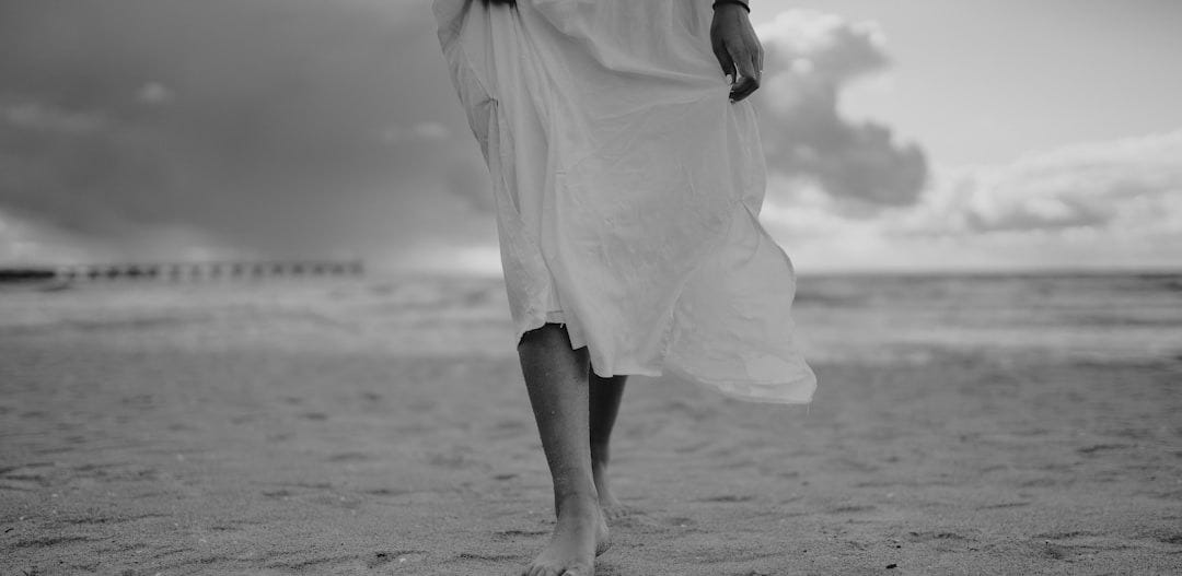 grayscale photo of woman in dress walking on beach