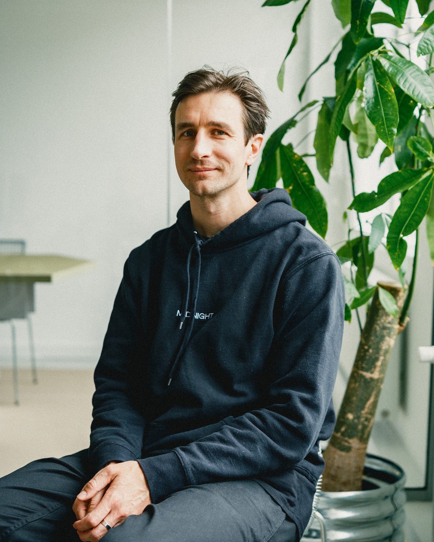 Photograph of a white man sitting down with a backdrop of plants and an office desk