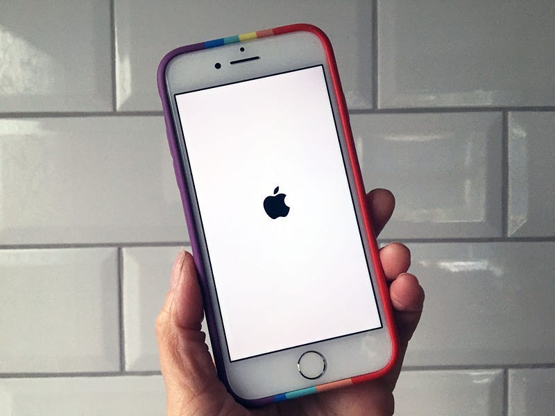 A hand holding an iPhone. The screen is showing the Apple icon of never-loading-again hell