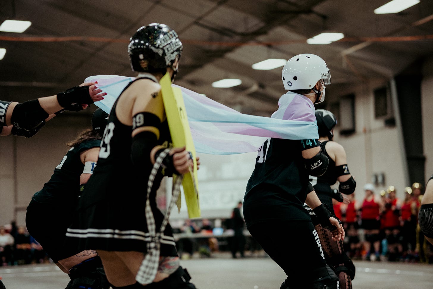 Emily, in their roller derby uniform and protective gear, including helmet and face mask, skates with a pack of fellow skaters, wearing a trans pride flag as a cape.