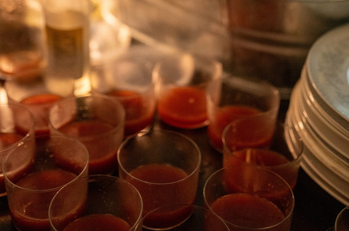 A row of small glass cups filled with bright red watermelon soda sitting on a counter next to a large galvanized metal ice tub and glass bottles.