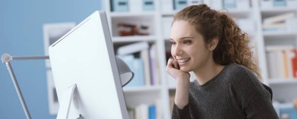 Young smiling beautiful woman using a computer in the office and connecting online, she is watching videos and films Young smiling beautiful woman using a computer in the office and connecting online, she is studying and working person smiling at computer screen stock pictures, royalty-free photos & images