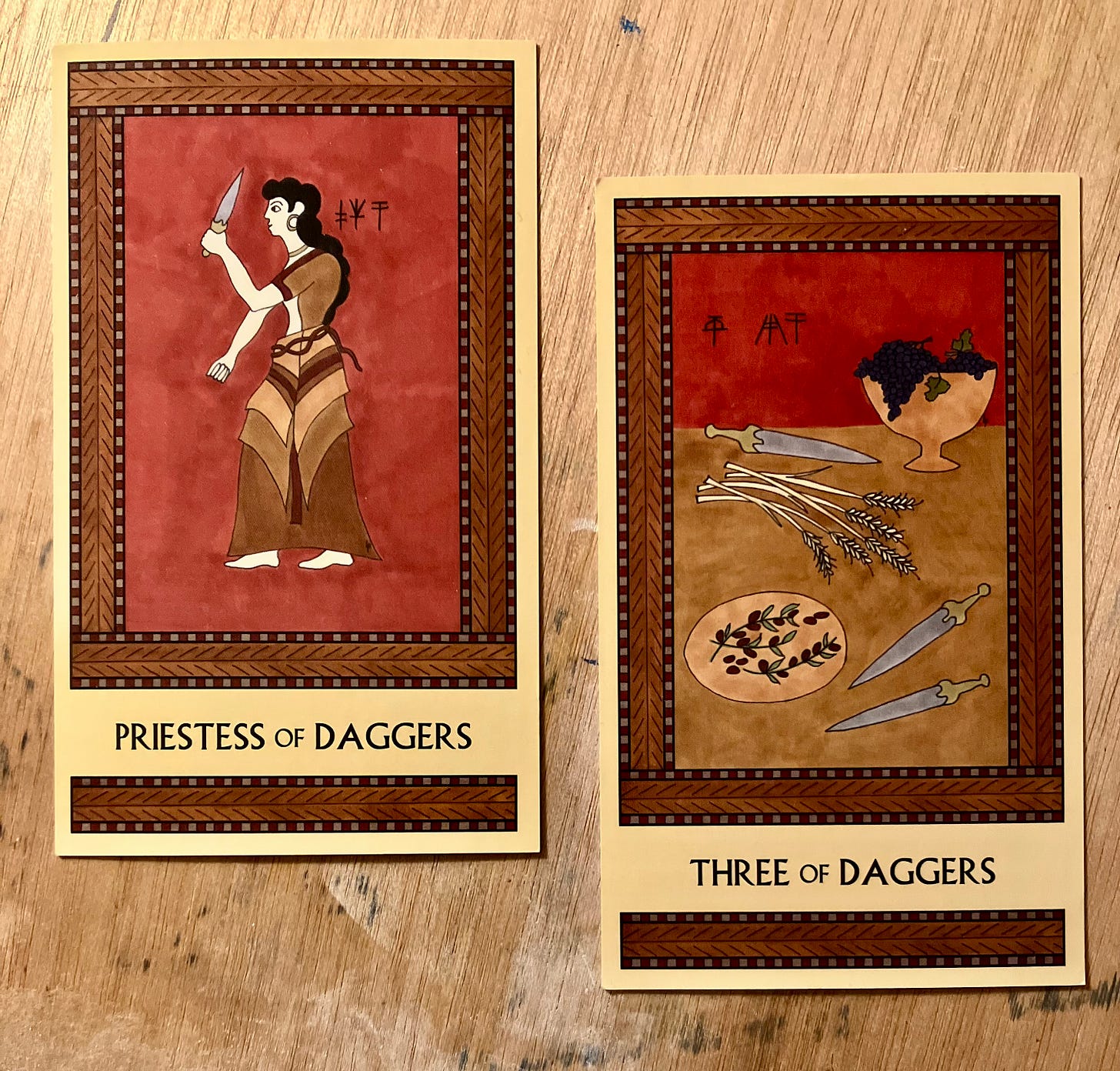 Two Minoan Tarot cards side by side on an ink-stained light wood surface (my art desk). Both cards are painted in shades of deep red, brown, and tan. The Priestess of Daggers depicts a Minoan woman with long hair and a long dress. She is standing, facing left, holding up a dagger in front of her. The Three of Daggers depicts a table laden with a footed bowl overflowing with grapes, a platter of olives, a pile of stalks of grain, and three daggers.