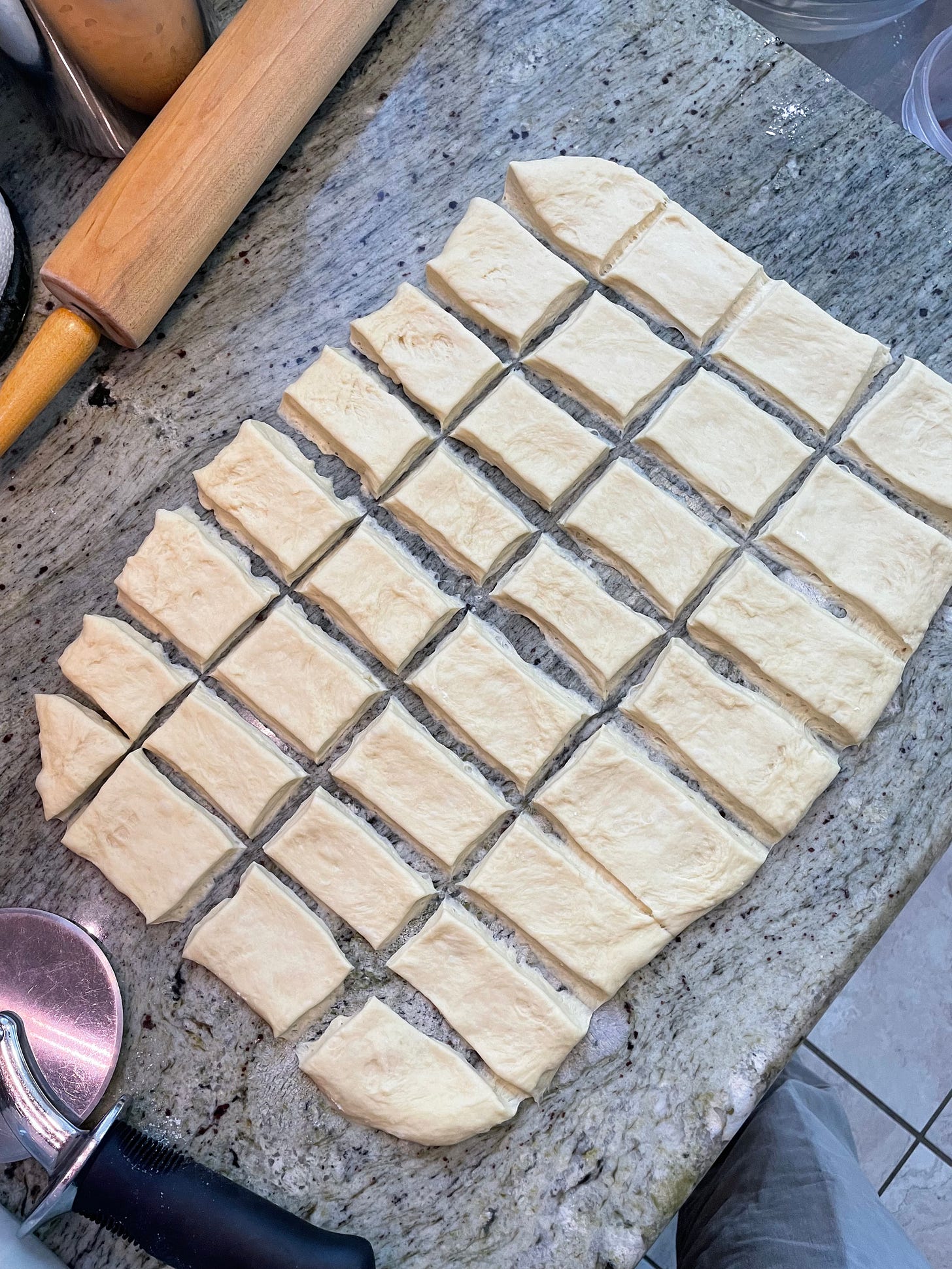 Light colored dough, thin and cut into 32 rectangles, on counter with rolling pin and pizza whieel cutter.