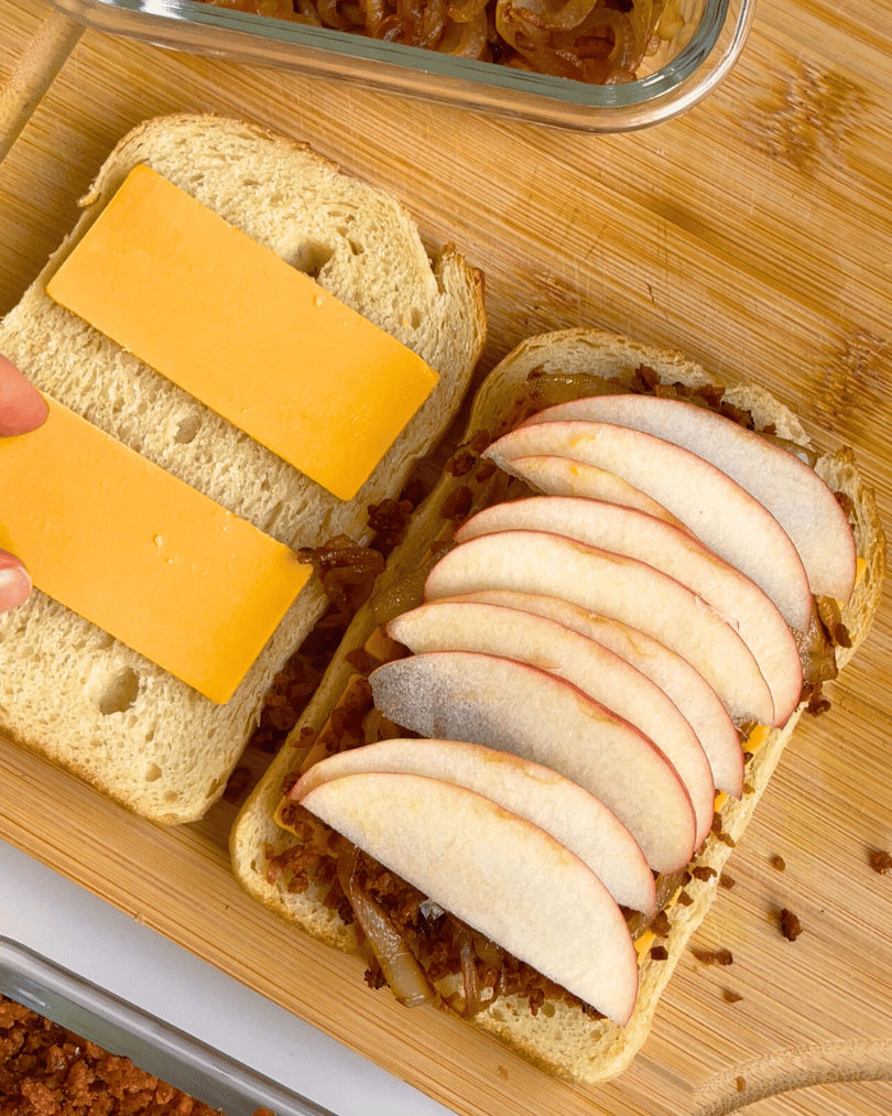 two slices sourdough bread with vegan cheddar on them and one side with vegan bacon bits, caramelized onions, and apple slices on it