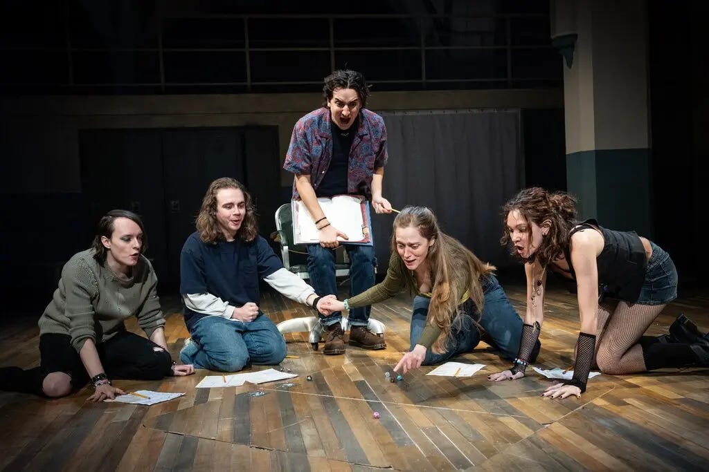 Four actors in casual wear sit on the floor playing a game, while a fifth actor, holding an open book, yells at the players. Four actors in casual wear sit on the floor playing a game, while a fifth actor, holding an open book, yells at the players.