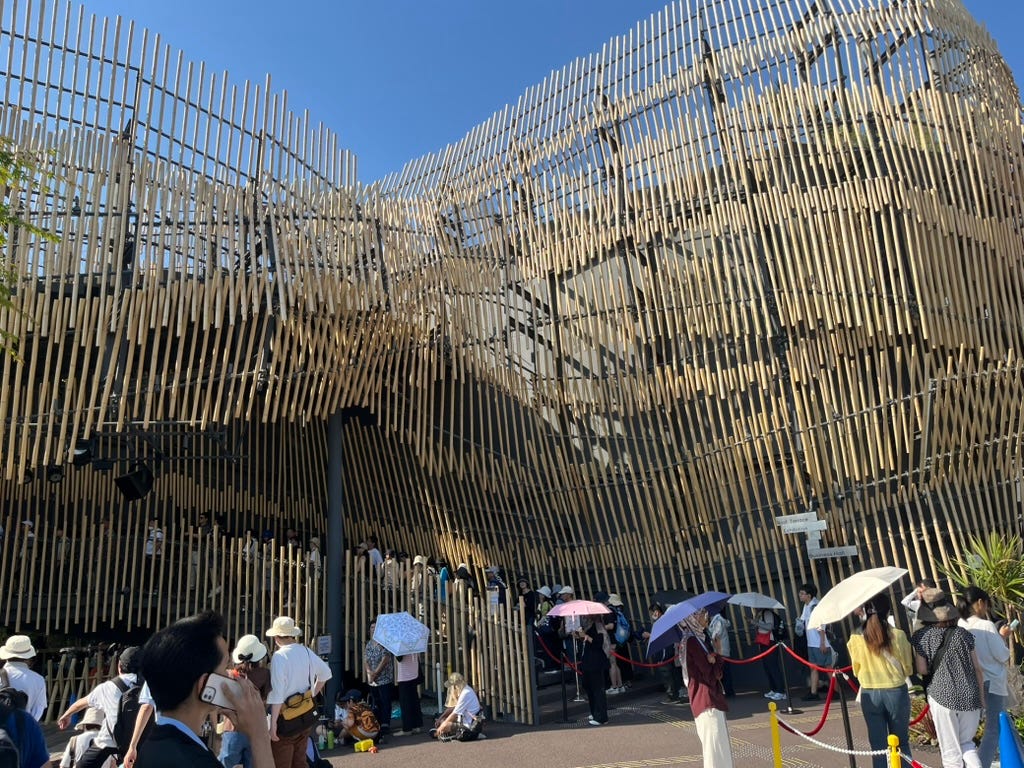 A curvy building with exterior made of yellowish bamboo. People are lining up on the stairs, and the line continues on the ground level.