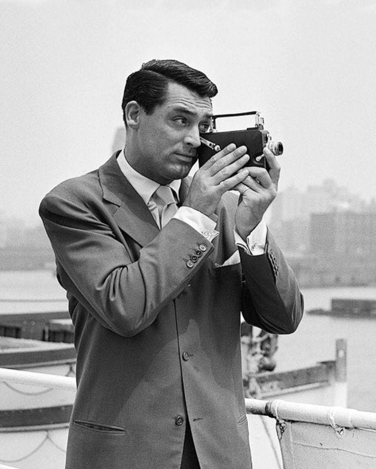 This may contain: a man in a suit holding a camera up to his face while standing on a boat This may contain: a man in a suit holding a camera up to his face while standing on a boat