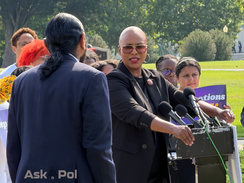 A Black female politician who's bald and in gold framed sunglasses speaks to a male African American politician on a sunny day at a press conference A Black female politician who's bald and in gold framed sunglasses speaks to a male African American politician on a sunny day at a press conference