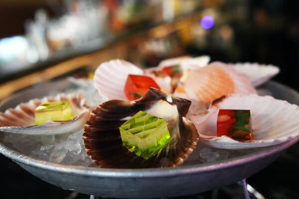 Cubes of yellow, green, and red jelly resting in seashells on a silver tray of ice. 