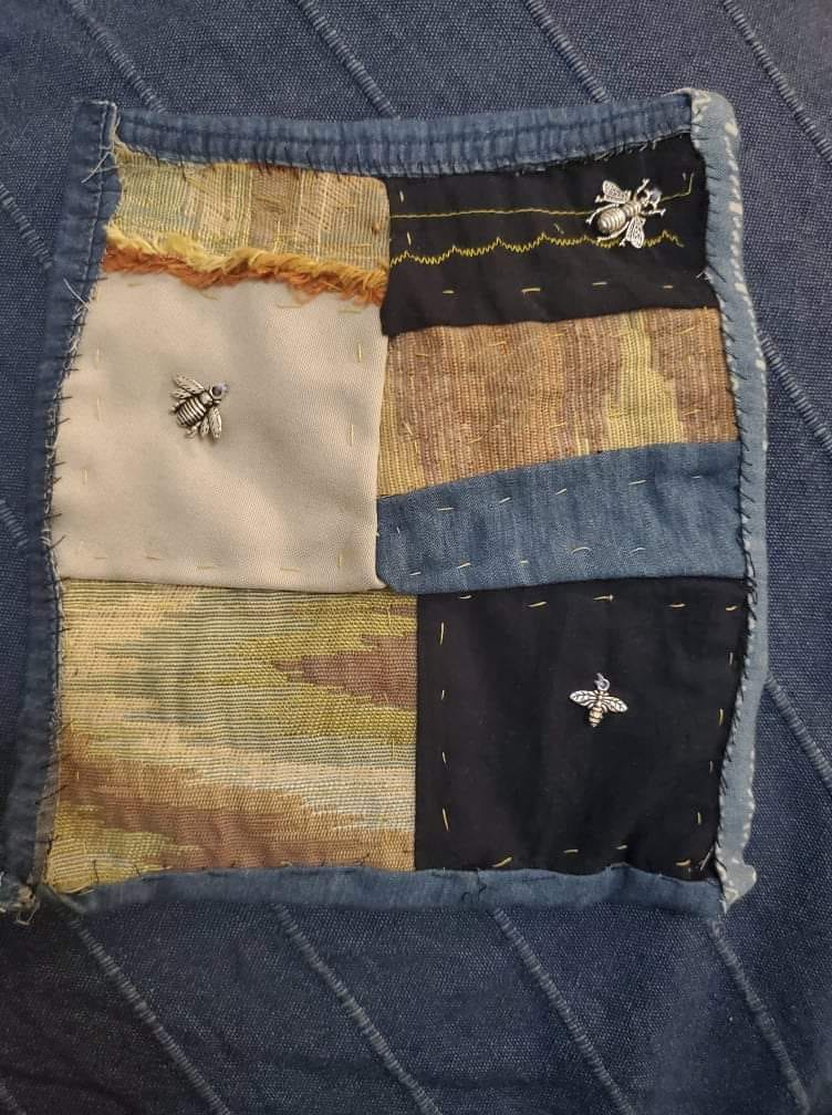 A quilt made with patches of beige, lavender, medium blue, and dark blue cloth is shown against a medium blue cloth background. The quilt features three small silver charms in the shape of honeybees. The charms are sewn onto various patches. 