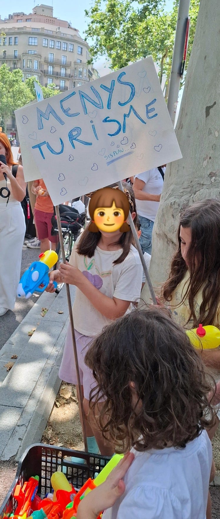 Una bambina tiene un cartello con la scritta "MENYS TURISME" (Meno turismo) scritto in maiuscolo blu con cuoricini disegnati intorno alle lettere. Indossa una maglietta bianca con una ciliegia rosa disegnata sopra e tiene in mano due pistole ad acqua, una blu e una gialla. Di fronte, si vede la parte posteriore della testa di un'altra bambina con capelli ricci e scuri che si china su un cestino pieno di pistole ad acqua colorate.