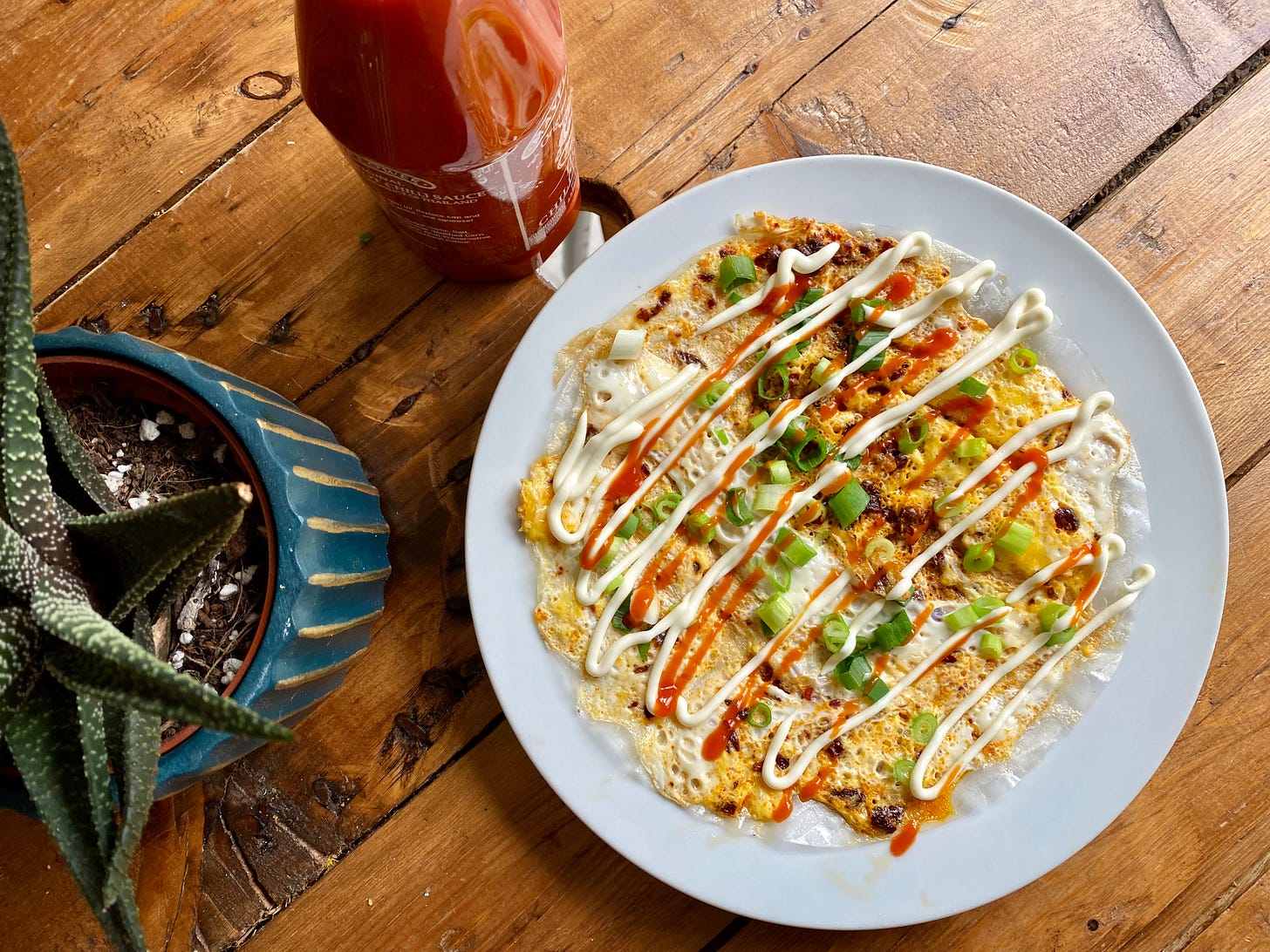 Plate with an omelette on top scattered with spring onions. A bottle of sriracha and a cactus are next to the plate. 