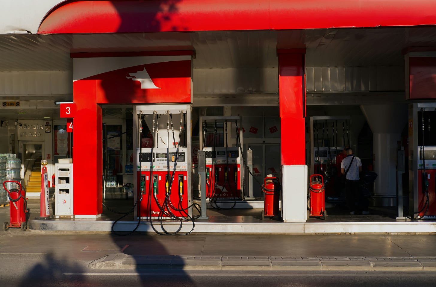 A brightly lit gas station island under a wide red and white canopy. Multiple red fuel dispensers with long black hoses stand on a raised concrete platform. A stylized white dog logo is visible on the prominent red pillar to the left. In the background on the right, a person stands near one of the pumps, while red fire extinguishers sit on wheels near the pillars. Shadows are cast across the foreground asphalt. A brightly lit gas station island under a wide red and white canopy. Multiple red fuel dispensers with long black hoses stand on a raised concrete platform. A stylized white dog logo is visible on the prominent red pillar to the left. In the background on the right, a person stands near one of the pumps, while red fire extinguishers sit on wheels near the pillars. Shadows are cast across the foreground asphalt.