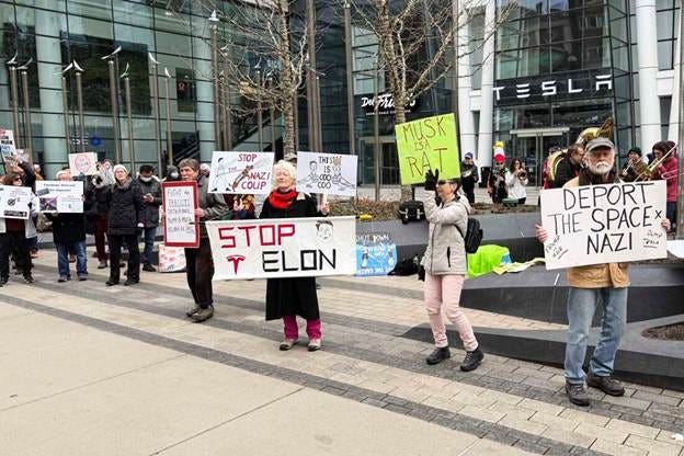 Protesters holding signs rally outside a Tesla store in Boston. One sign says "Stop Elon." 