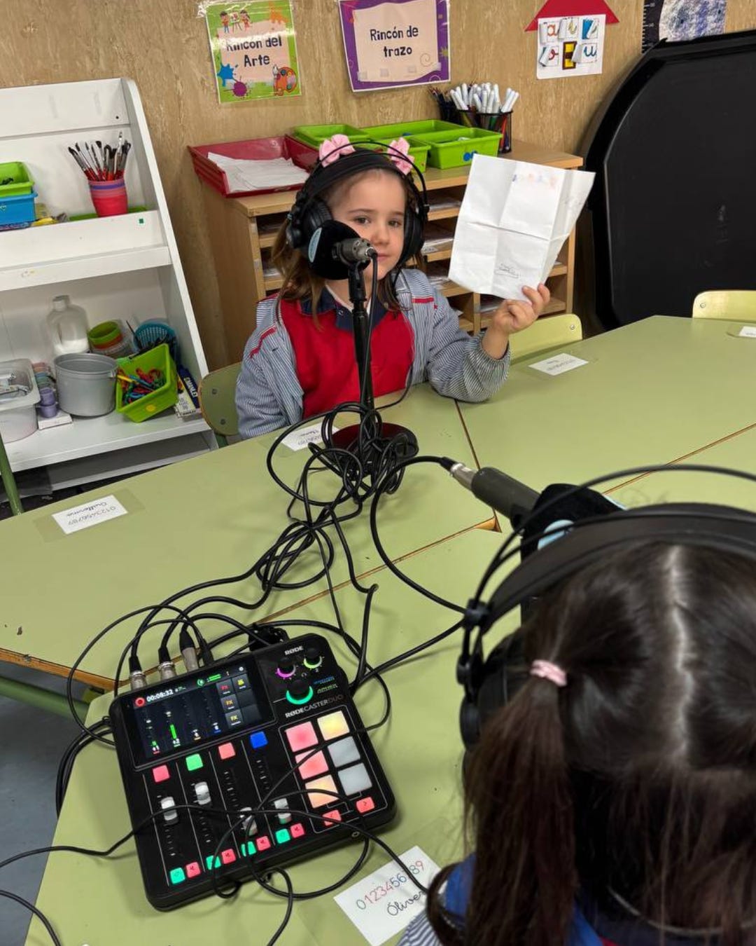Niña con auriculares grabando en un micrófono en una mesa de aula mientras sostiene una hoja de papel. Frente a ella se ve otra niña también con auriculares y una mesa de mezcla RØDECaster conectada a los micrófonos. Al fondo aparecen estanterías con material escolar y carteles de actividades como “Rincón del arte” y “Rincón de trazo”. Niña con auriculares grabando en un micrófono en una mesa de aula mientras sostiene una hoja de papel. Frente a ella se ve otra niña también con auriculares y una mesa de mezcla RØDECaster conectada a los micrófonos. Al fondo aparecen estanterías con material escolar y carteles de actividades como “Rincón del arte” y “Rincón de trazo”.