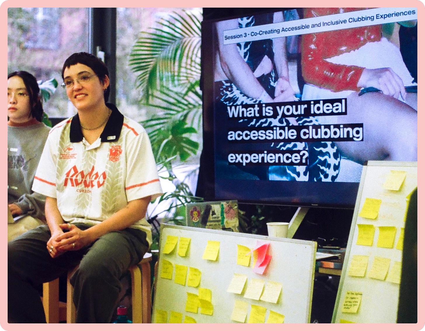 A photo of Ali sitting on a stool and looking off camera, smiling. Beside her, loads of yellow post-its have been stuck to a white board, and a large digital screen reads "What is your dream accessible clubbing experience?"