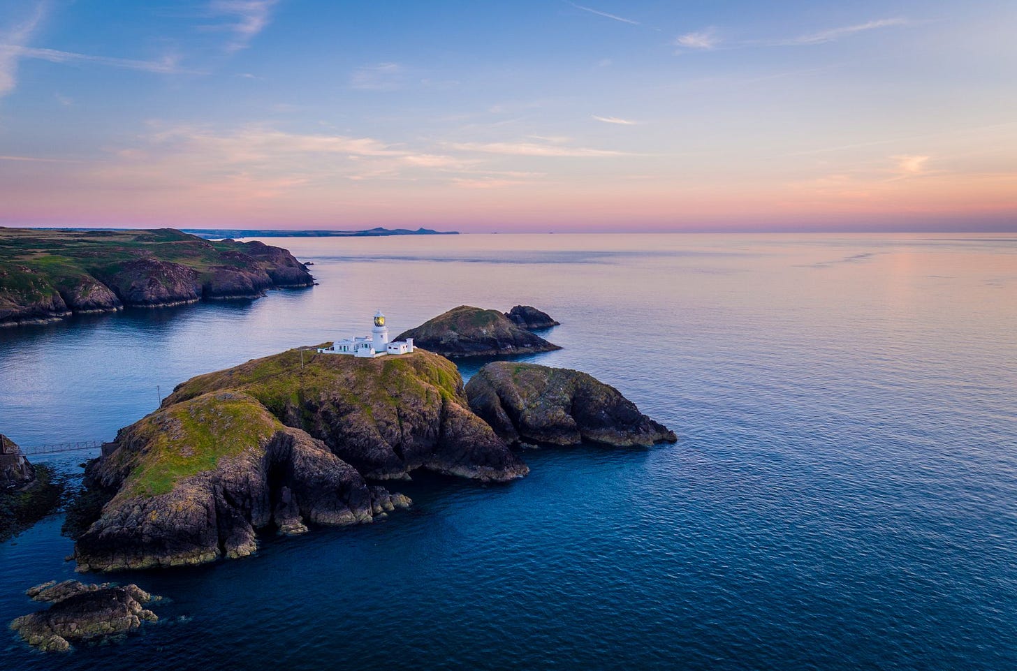 An aerial view of Strumble Head Lighthouse at dusk. The white lighthouse complex is perched on the small, rugged, grass-topped island of Ynys Meicel, surrounded by deep blue sea under a soft pink and purple sky.