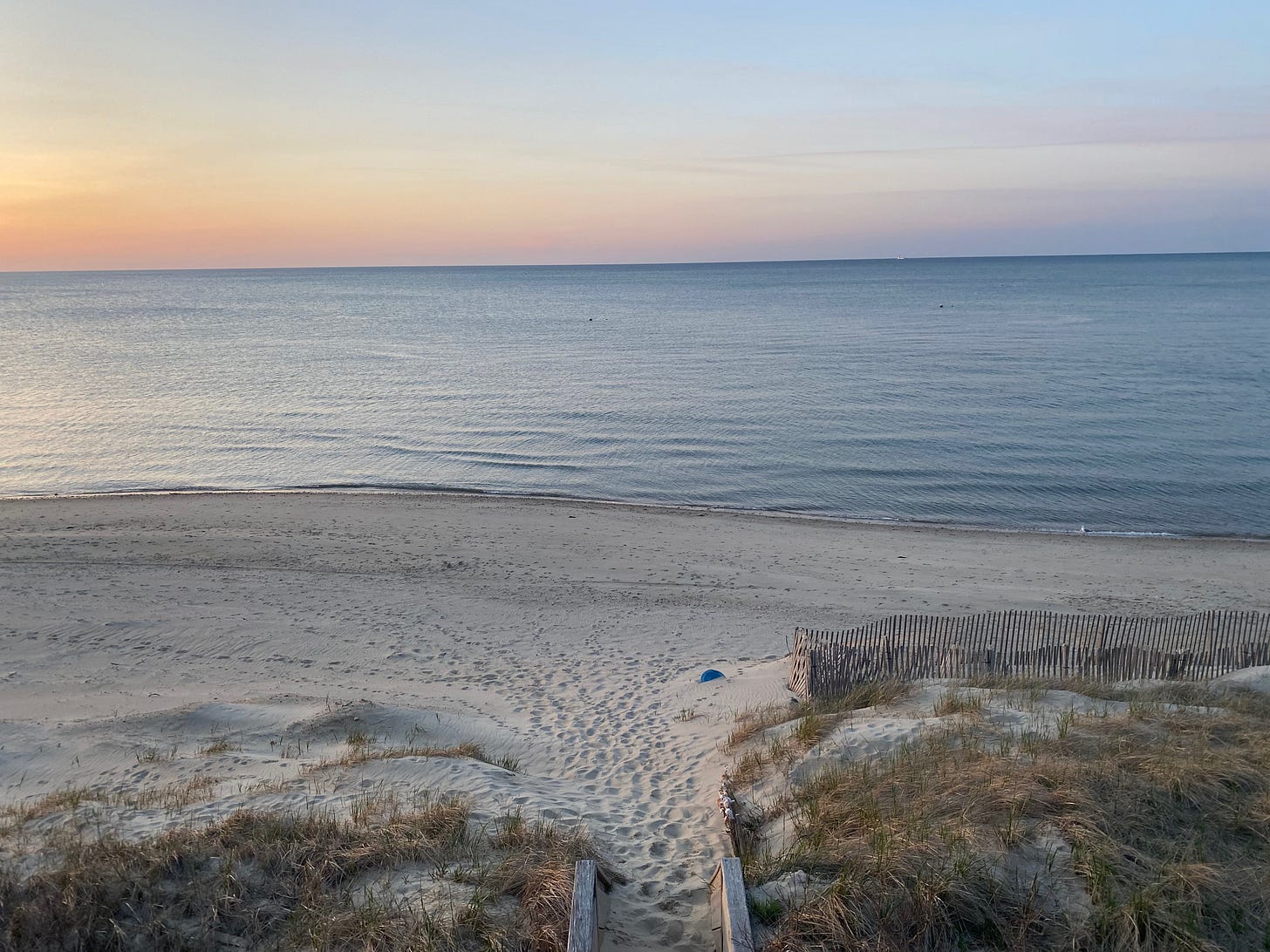 The beach at sunset. The water is pale blue and still; the sky a mix of blue, orange, pink and purple. The sand is covered in footprints.