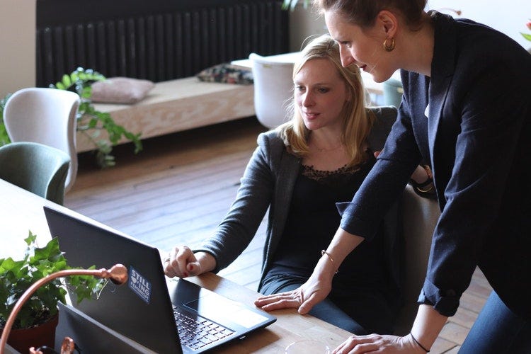 A woman is leaning over a desk helping another woman with something on her computer