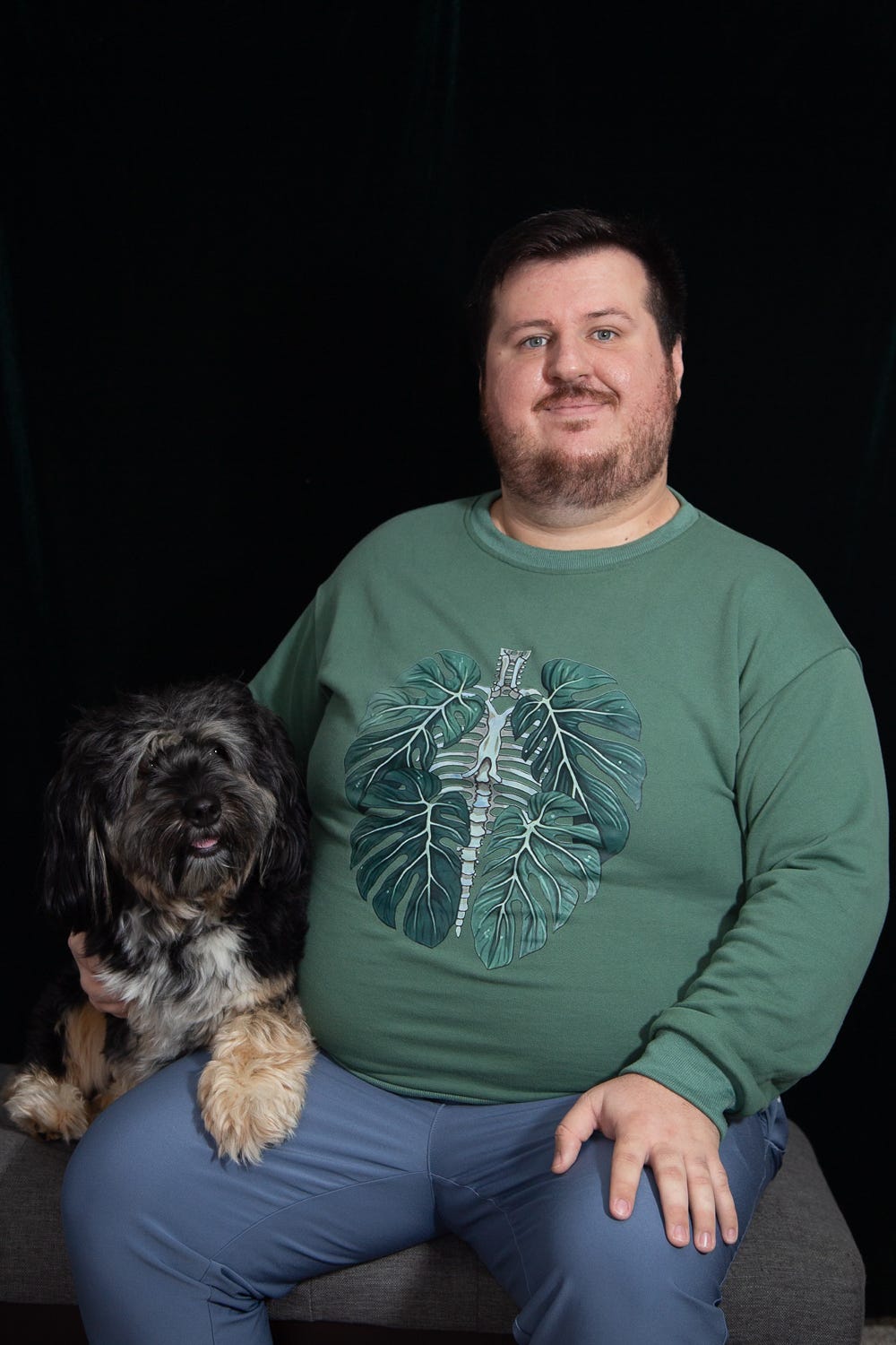 A portrait of Jordan and his dog, Arlo. They are sitting on a bench with a black backdrop. Jordan is wearing a sage green sweatshirt with an image of a ribcage with leaves surrounding it and blue pants. Jordan has white skin, short brown hair, a beard, moustache and blue eyes. Arlo is sitting next to him, with his paw in Jordan’s lap. Arlo is a small dog, with long hair, floppy ears and black and beige coloring. A portrait of Jordan and his dog, Arlo. They are sitting on a bench with a black backdrop. Jordan is wearing a sage green sweatshirt with an image of a ribcage with leaves surrounding it and blue pants. Jordan has white skin, short brown hair, a beard, moustache and blue eyes. Arlo is sitting next to him, with his paw in Jordan’s lap. Arlo is a small dog, with long hair, floppy ears and black and beige coloring.