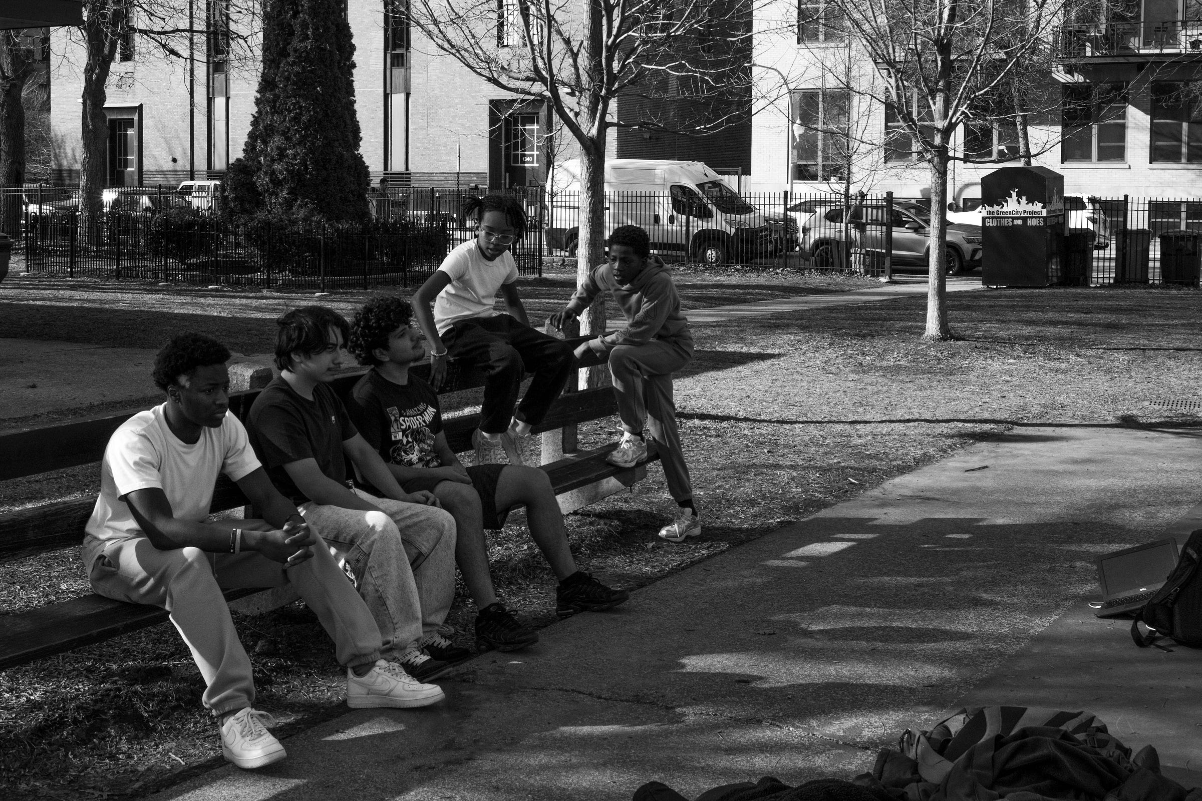 A black-and-white photograph of a group of young men sitting and leaning on a park bench, engaged in conversation. Two of them sit on the top of the bench, one gesturing as if mid-sentence, while the others listen attentively. The background features a fenced-in urban park with bare trees, a clothing donation bin, parked cars, and an apartment building. Sunlight filters through the trees, casting long shadows on the pavement, emphasizing the relaxed yet engaged atmosphere.