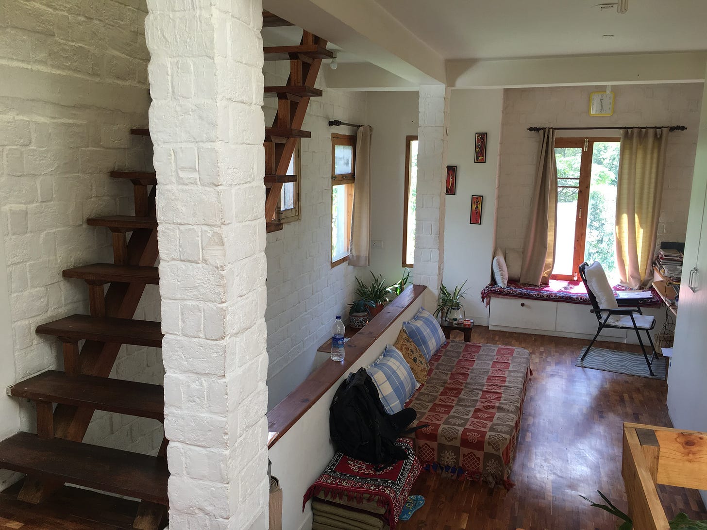 A white brick interior with a brown wooden staircase to the left side. Wooden floors and furniture are shown, covered in red patterned fabrics.