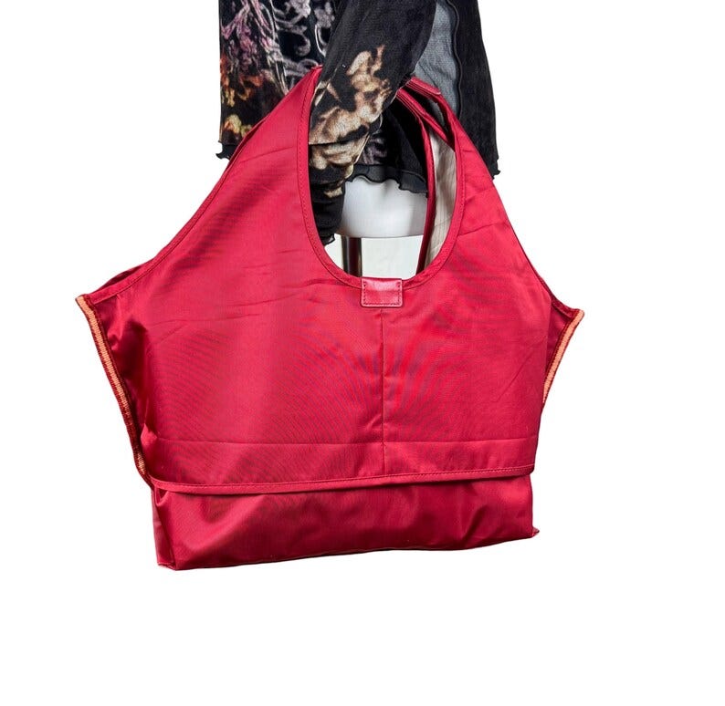 May include: A large, vibrant red tote bag made of a smooth, synthetic material. The bag features a wide, integrated handle design, a horizontal seam across the lower section, and a rectangular tab detail at the center of the top opening.