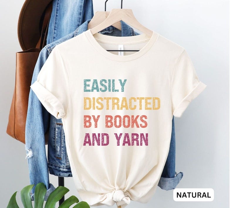 May include: A natural colored t-shirt with the text 'EASILY DISTRACTED BY BOOKS AND YARN' in rainbow colors. The text is in a vintage style font and has a distressed look.