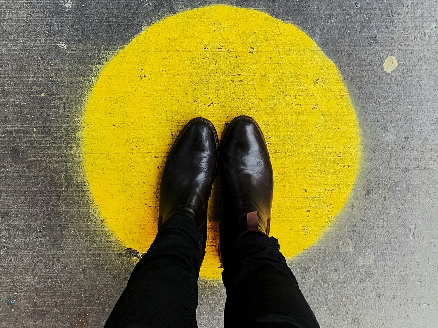 Someone in black throusers and leather shoes stands in a bright yellow, sunlike chalk circle on grey paving.