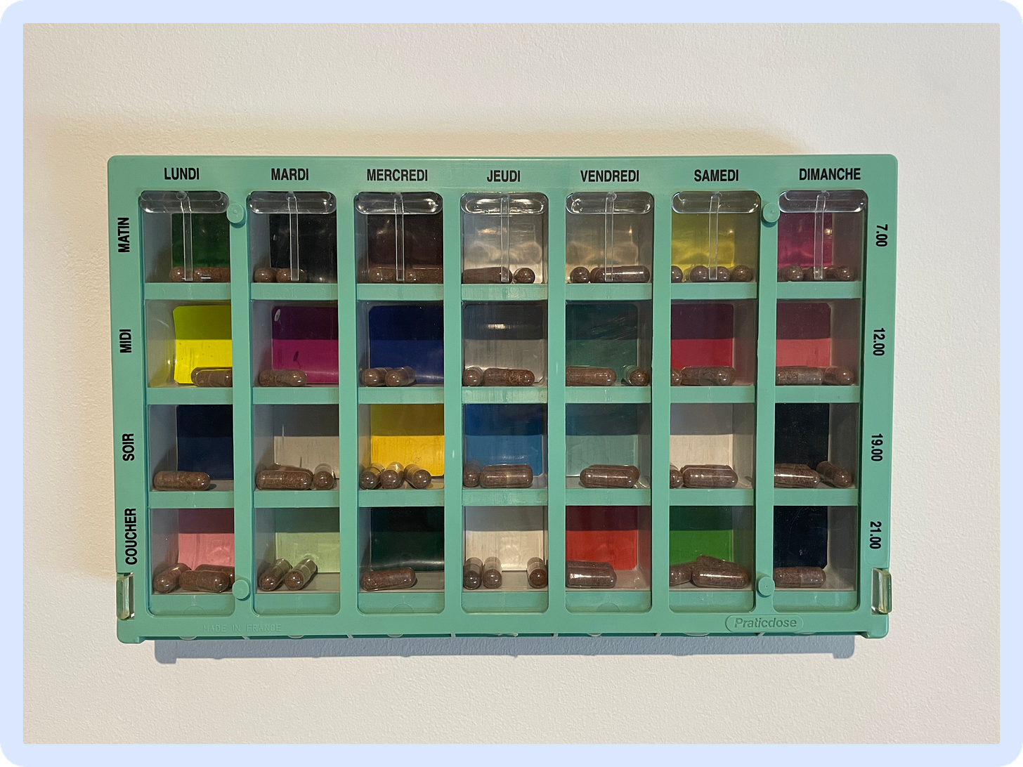 A photo of a green plastic pill divider, with the days of the week in French in black horizontal text, and the times of the day also in black and in French ("voucher", "sour", "midi", "matin") vertically. Inside, clear capsules are filled with dark brown powder, and the individual pill compartments are backed with different bright coloured paper (eg. pink, teal, sage green or bright yellow, among others). A photo of a green plastic pill divider, with the days of the week in French in black horizontal text, and the times of the day also in black and in French ("voucher", "sour", "midi", "matin") vertically. Inside, clear capsules are filled with dark brown powder, and the individual pill compartments are backed with different bright coloured paper (eg. pink, teal, sage green or bright yellow, among others).