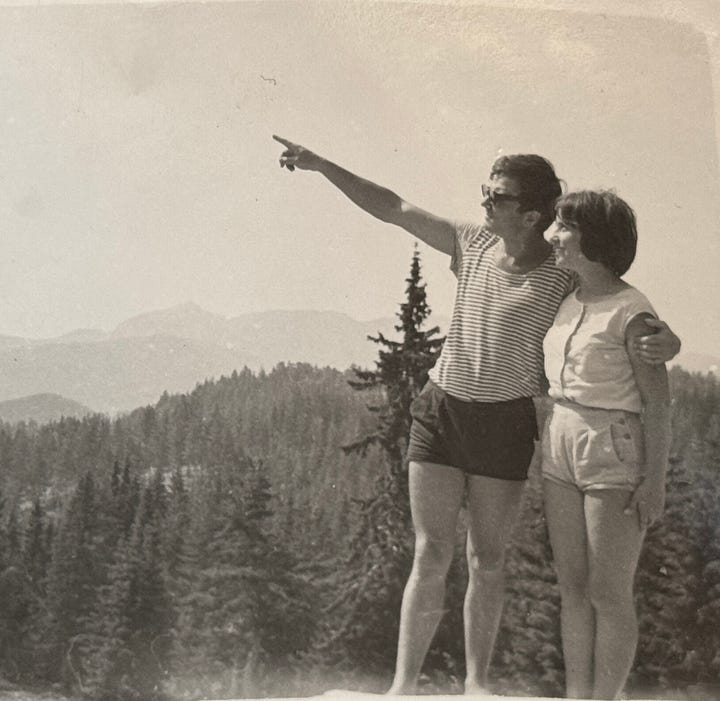 A black-and-white photo of a young couple in the late 1960s standing outdoors on a scenic hillside. The man, wearing sunglasses and a striped t-shirt with shorts, has his arm around the woman and is pointing toward the distance. The woman, wearing a sleeveless buttoned shirt and shorts, is smiling as she looks in the same direction. Tall evergreen trees and mountain peaks are visible in the background under a hazy sky.