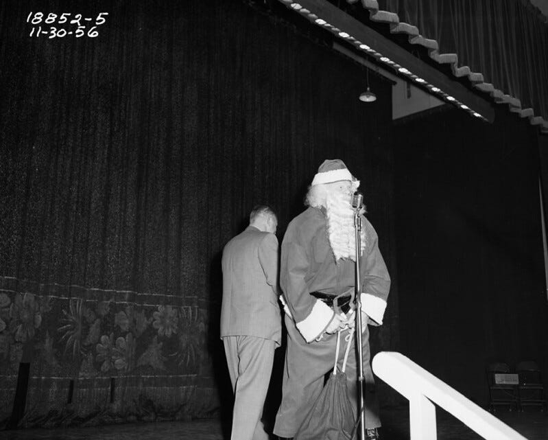 Black and white photo of Santa Clause on a stage holding a large gift bag