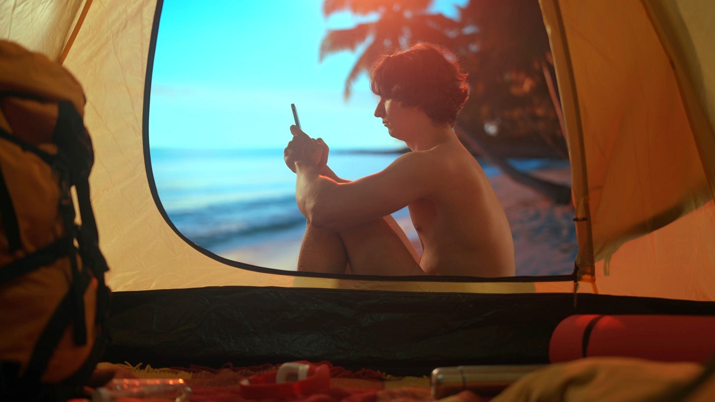 A nude person sits at the opening of a camping tent on a beach, looking at a smartphone, with the ocean and a palm tree visible in the background. A nude person sits at the opening of a camping tent on a beach, looking at a smartphone, with the ocean and a palm tree visible in the background.