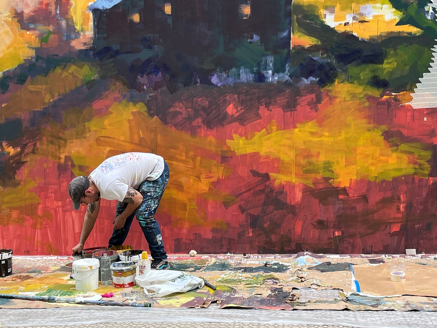 man bent over paints with a wash of color on wall behind him.
