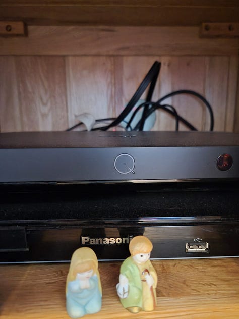 Small figures from a nativity scene in front of a model bakery and a DVD player. A small pottery sheep is sitting on top of a barn.