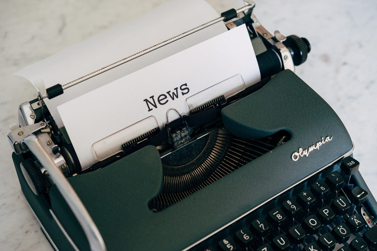 a green Olympia typewriter sits on a marble desk top. In the typewriter is a piece of paper with News typed on it.
