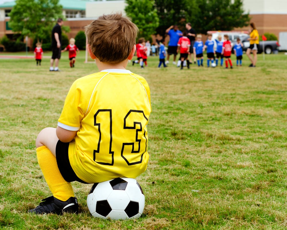 Young boy sitting on a soccer ball on the sideline alone