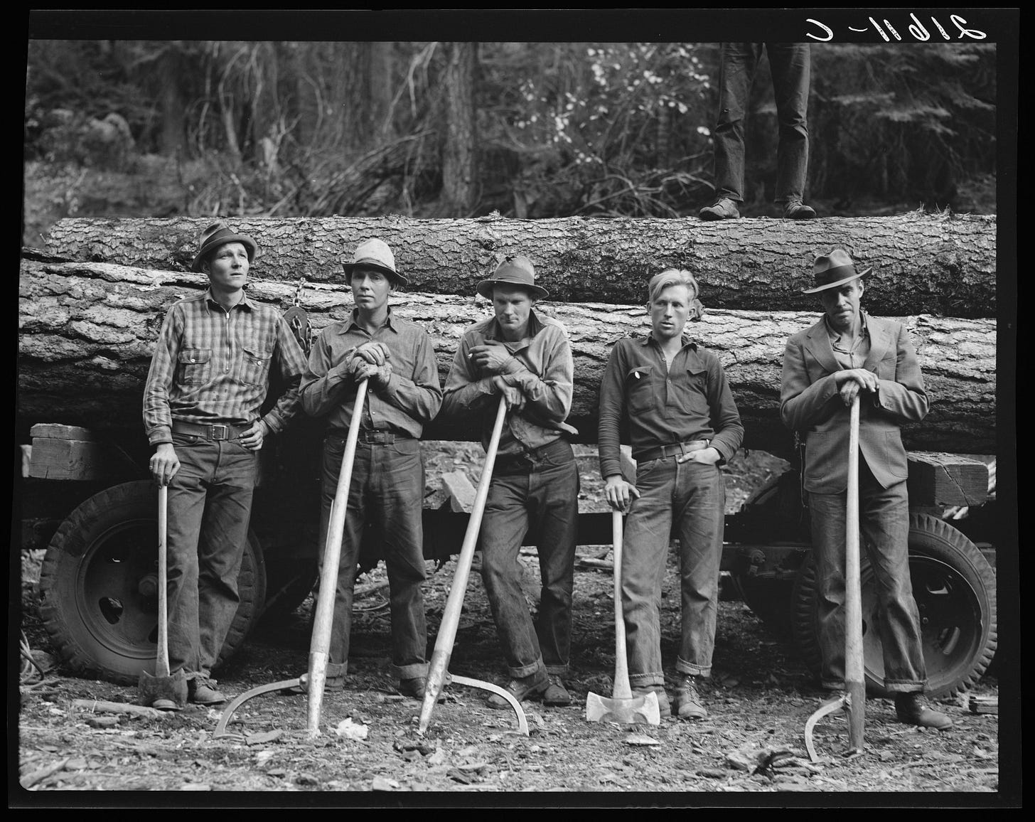 Five men stand in front of vehicle holding large tree truns. Each man rests his hand or hands on an upright mill tool.
