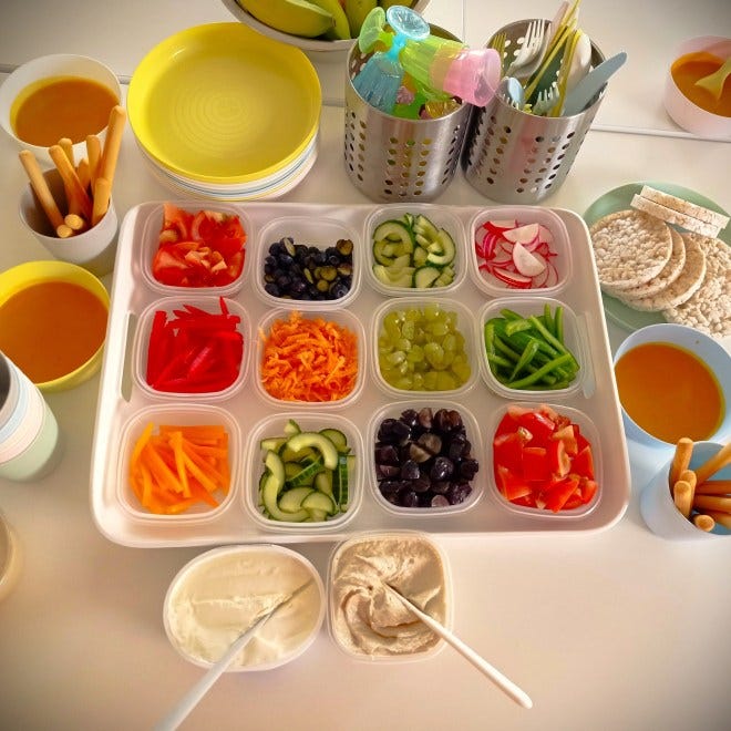 a tray with plastic tubs full of chopped fruit and veg with some plastic plates, cups and cutlery