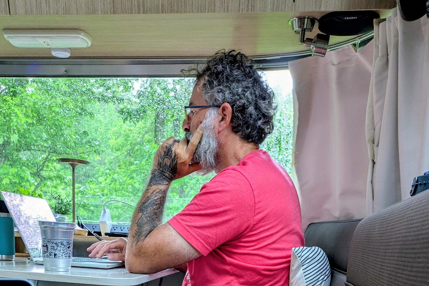 The author, a man with curly gray hair, a white beard, glasses, and tattooed arms, sits in an RV working at a laptop. He rests his chin on his hand, gazing out a large window at green trees, lost in thought. A desk lamp and coffee cup sit nearby. The author, a man with curly gray hair, a white beard, glasses, and tattooed arms, sits in an RV working at a laptop. He rests his chin on his hand, gazing out a large window at green trees, lost in thought. A desk lamp and coffee cup sit nearby.