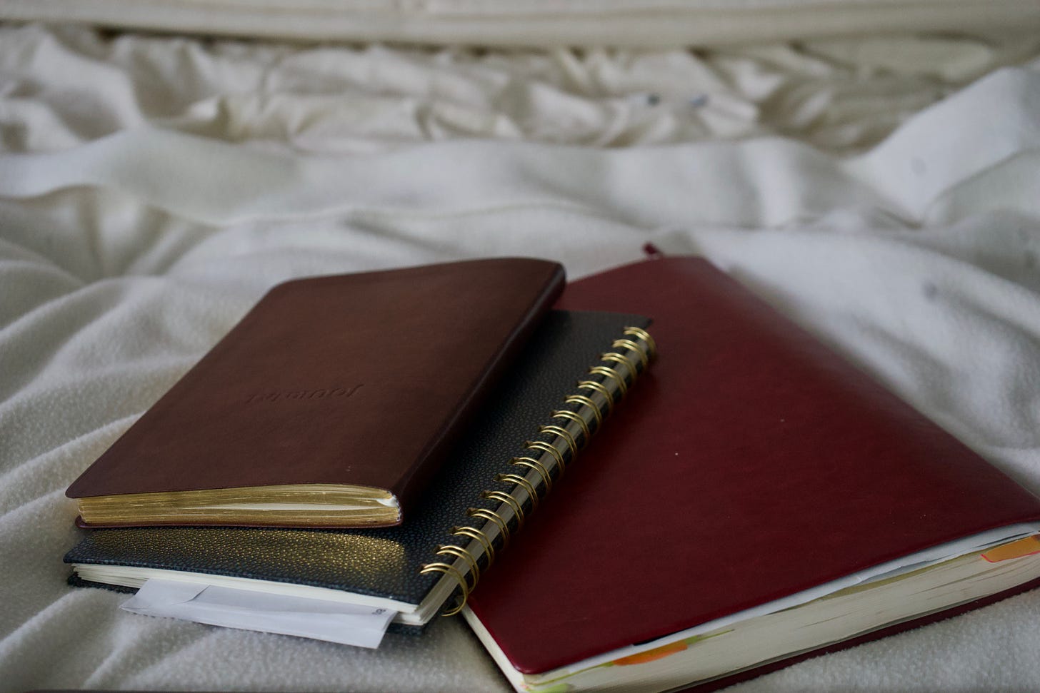 Stacked leather journals in warm brown and deep red rest on textured white bedding, shot at a low angle with shallow depth of field, highlighting worn edges, gold spiral binding, and the tactile weight of repetition. Stacked leather journals in warm brown and deep red rest on textured white bedding, shot at a low angle with shallow depth of field, highlighting worn edges, gold spiral binding, and the tactile weight of repetition.