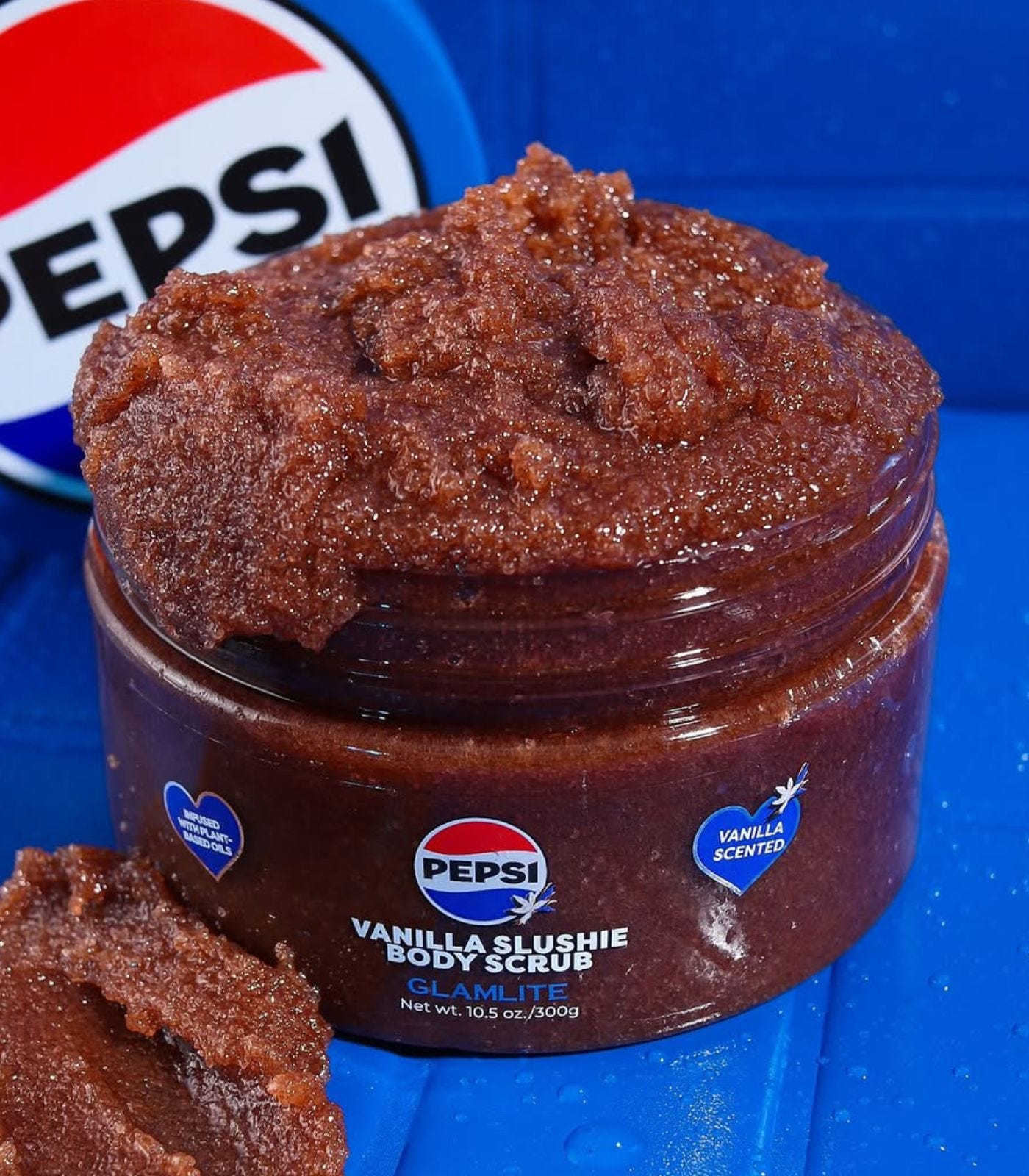 A tub of body scrub in front of blue tiles; it looks like its full of glittery diarrhea