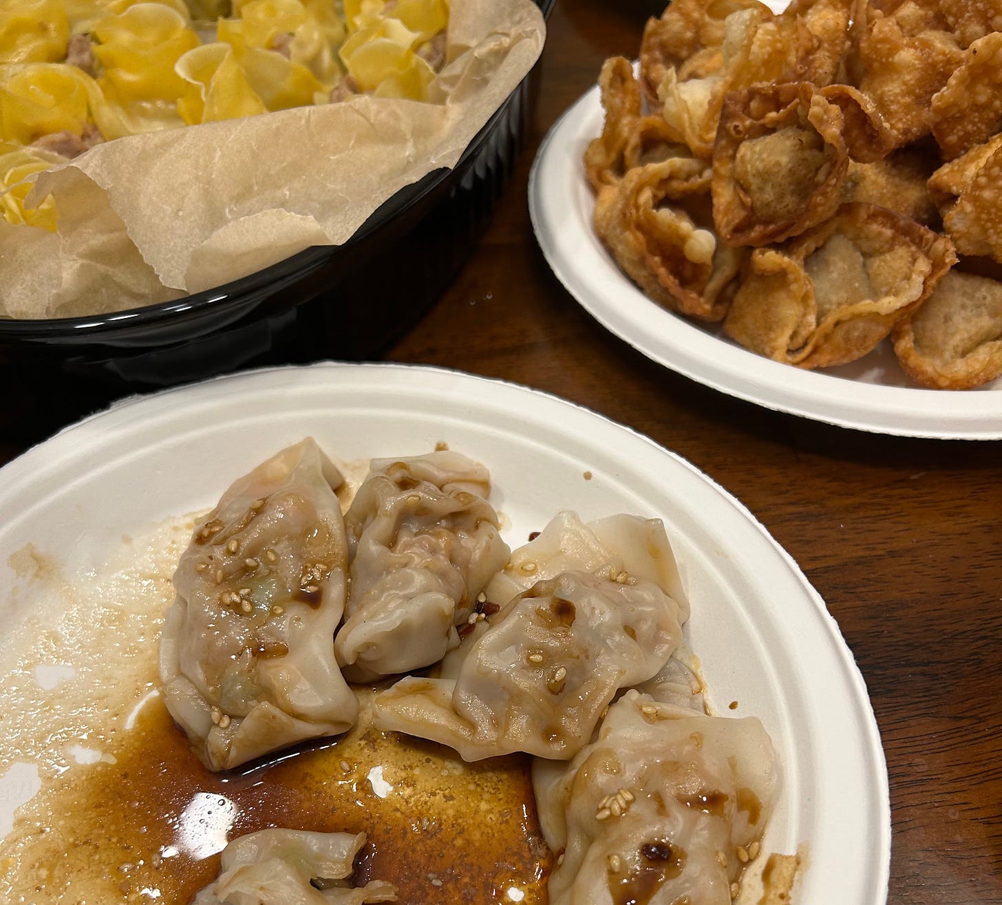 Three dishes of dumplings, yellow siu mai in the top left corner, deep-fried wontons in the top right corner, and boiled mandu in a soy sauce mixture in the foreground.